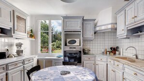 Una cocina con muebles blancos, una mesa de comedor con mantel azul y blanco, y vista a zonas verdes al exterior.