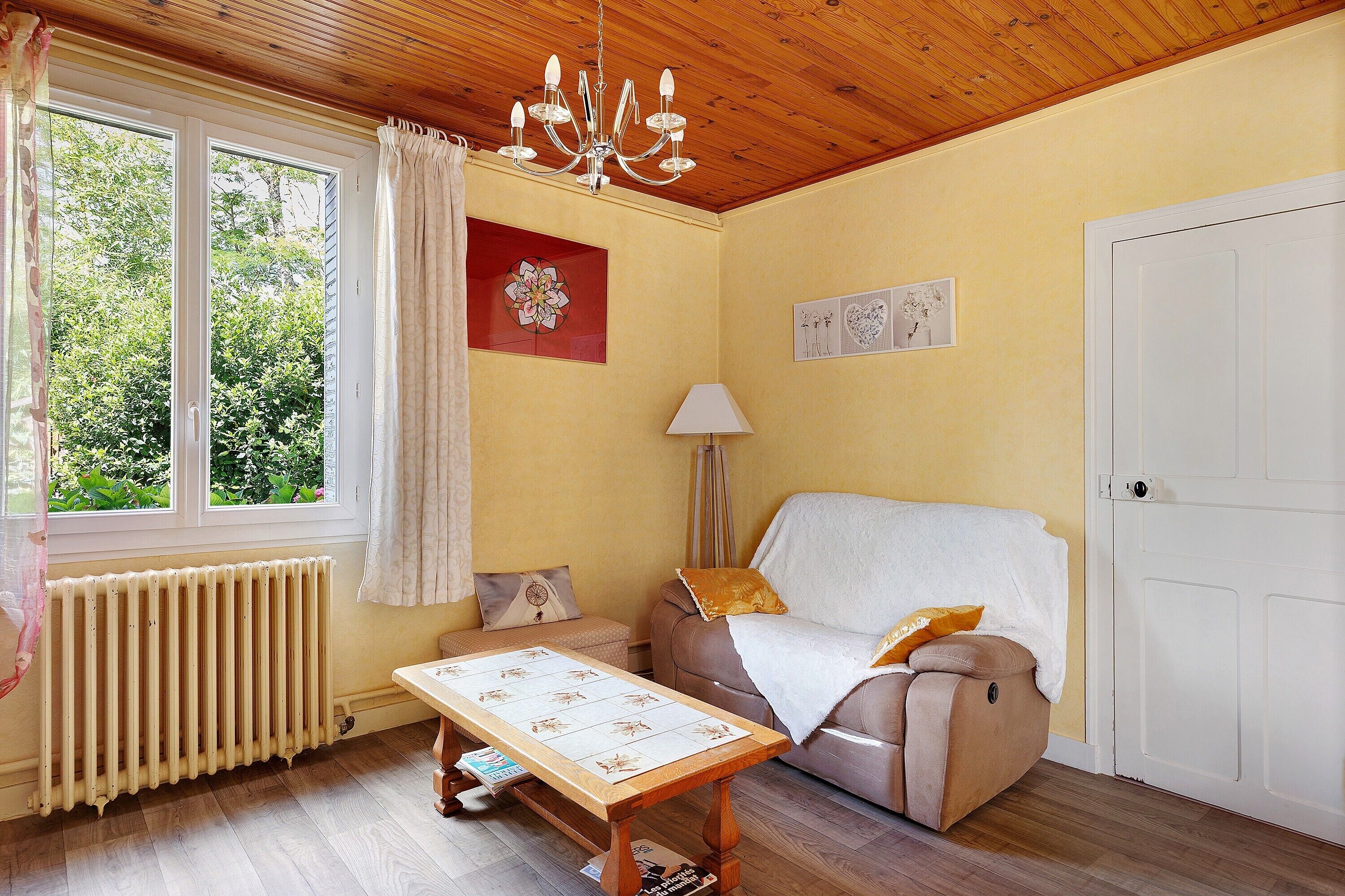 A cozy room with a wooden ceiling, a radiator, a sofa, a table with a floral tablecloth, and artwork on the walls.