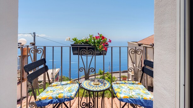 Uma varanda com mesa e cadeiras, um vaso suspenso com flores e vista para o mar.