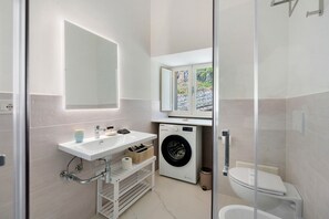 Une salle de bain moderne équipée d’un lavabo, d’un miroir, d’un lave-linge, de toilettes et d’une douche à parois de verre.