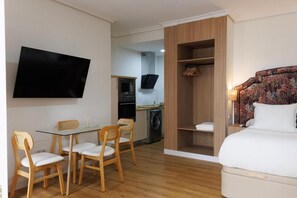 Une chambre d’hôtel compacte comprenant un lit, une télévision, un coin repas avec des chaises, une armoire en bois et un lave-linge à l’arrière-plan.