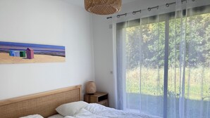 Ein Schlafzimmer mit Bett, Nachttisch, Lampe, einem Gemälde und einem großen Fenster mit Blick ins Grüne.