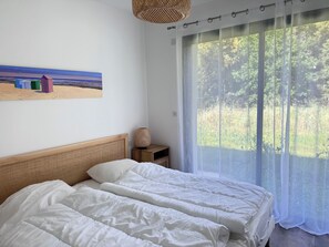Un dormitorio con cama, mesita de noche, lámpara, un cuadro y un ventanal con vistas al verdor.