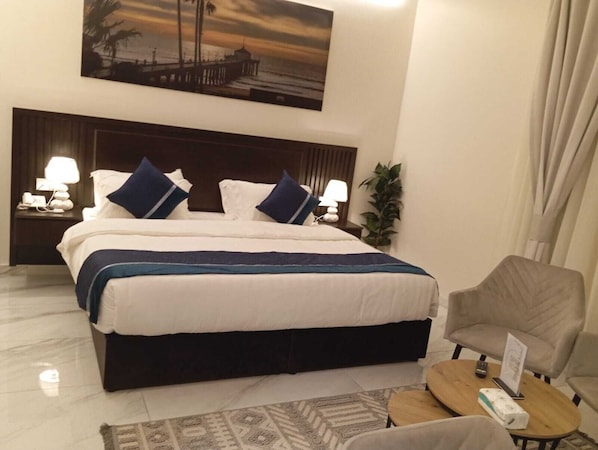 Habitación de hotel con una cama grande, dos mesitas de noche con lámparas, una silla y una pequeña mesa con un control remoto.