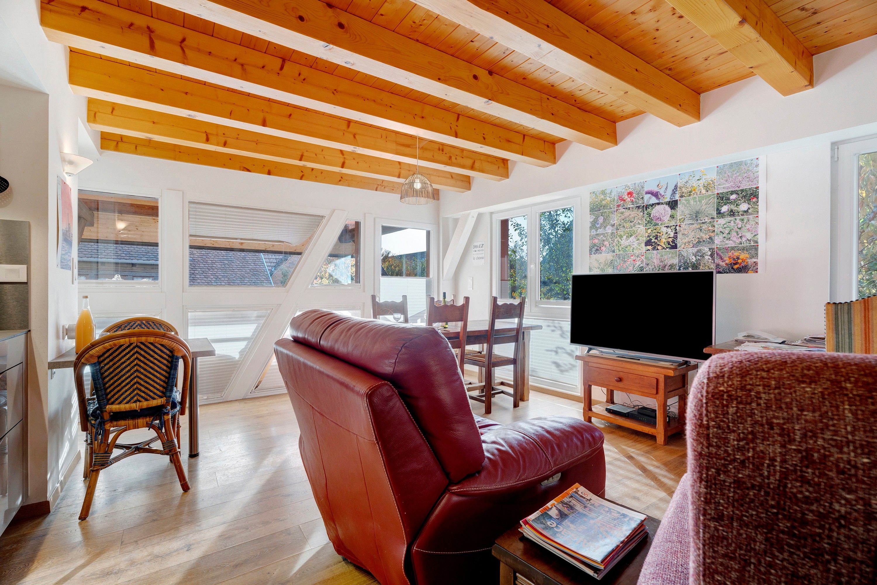 A living room with a red leather sofa, wooden flooring, a television, and a wall with a floral photo collage.