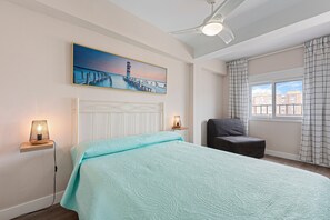 A bedroom with a bed, a nightstand with a lamp, a chair, a painting of a pier, and a window with curtains. - Apartment 'Málaga Beach, Costa del Sol' with Mountain Views, Wi-Fi & Air Conditioning (Málaga)