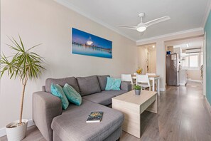 A modern living room with a grey sectional sofa, a wooden coffee table, a potted plant, and a painting of a sunset over a pier. - Apartment 'Málaga Beach, Costa del Sol' with Mountain Views, Wi-Fi & Air Conditioning (Málaga)