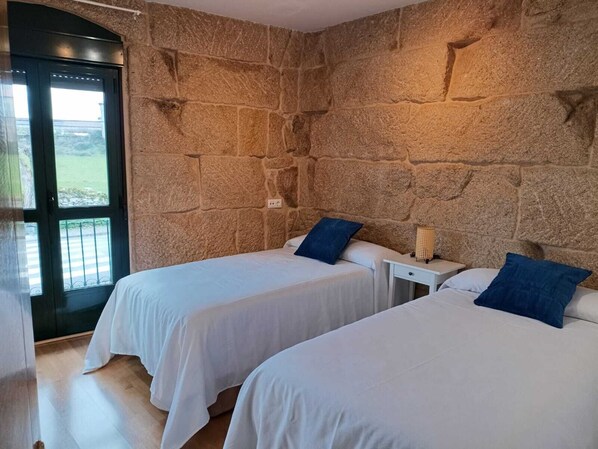 Una habitación con dos camas, paredes de piedra, una puerta que da al exterior y una pequeña mesa con una lámpara.