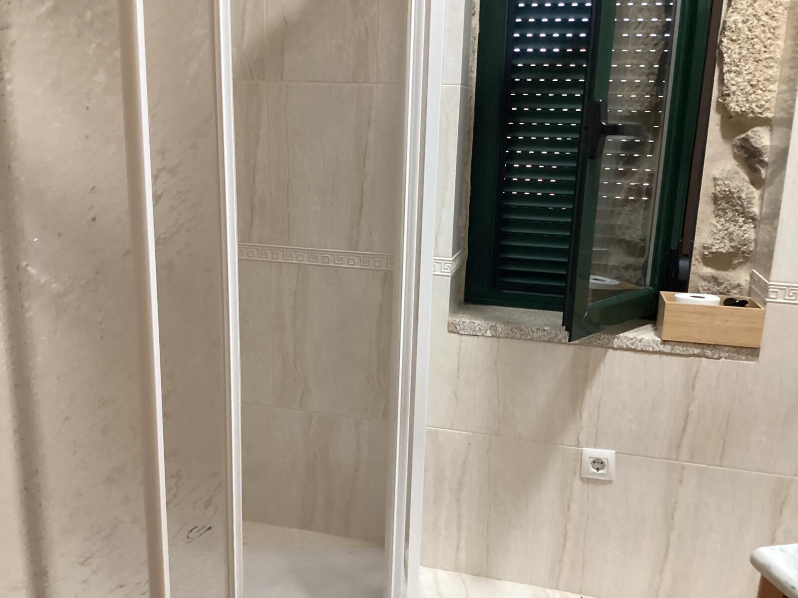A bathroom with a shower enclosure, a window with shutters, and a small shelf with toiletries.