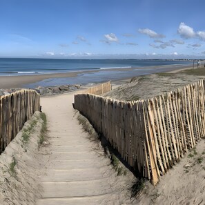 Ein sandiger Küstenpfad führt zum Meer, begleitet von einem Holzzaun und mit freiem Blick auf das Meer und den Himmel.