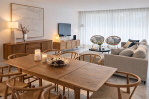 Ein modernes Wohnzimmer mit einem Holzes Esstisch, zwei Korbstühlen, einem Sofa und einem Fernseher an der Wand.
