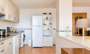 A compact kitchen with a washing machine, a white refrigerator, and a built-in shelf unit.