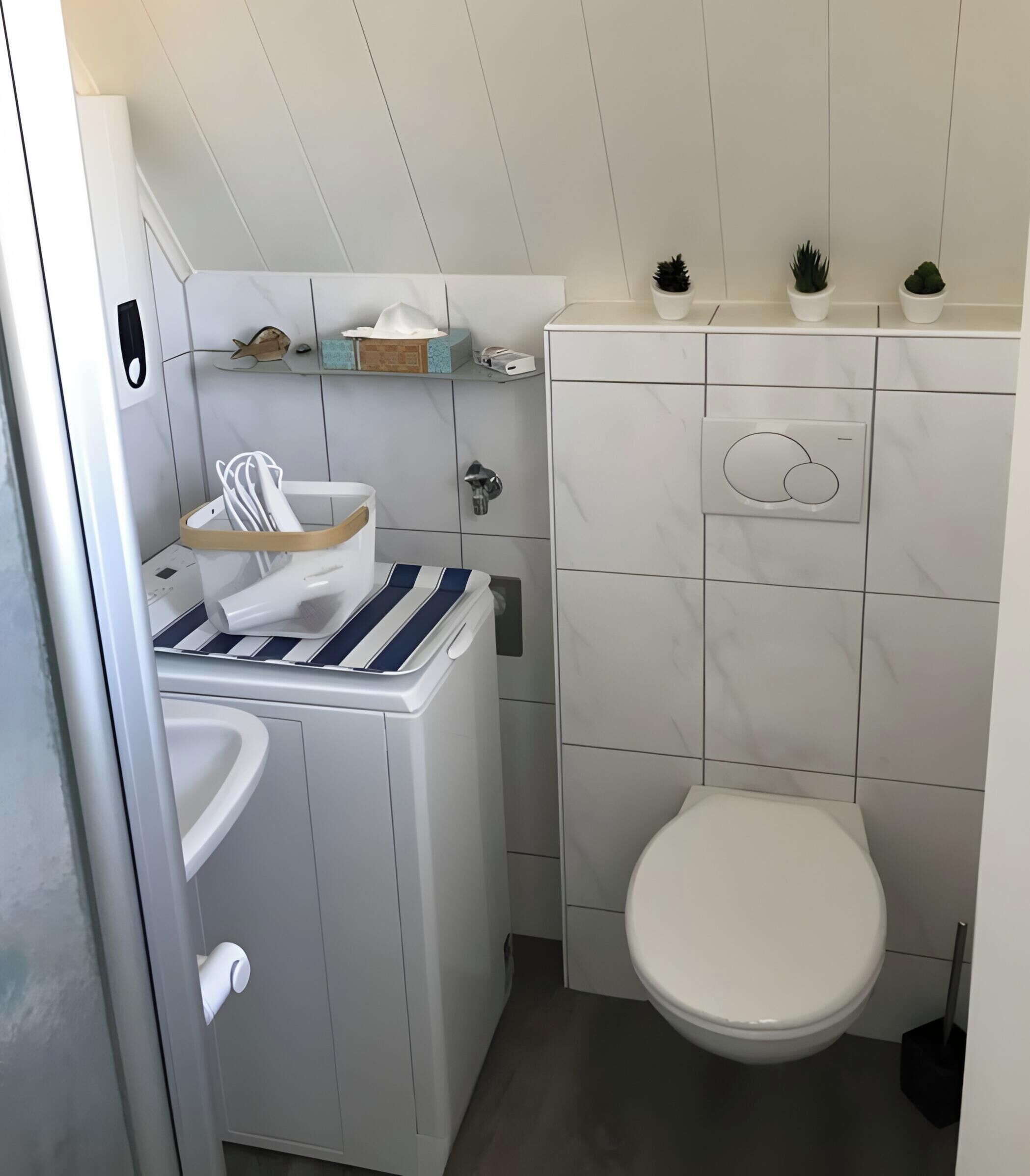 A compact bathroom with a toilet, a washing machine, and a small shelf with toiletries.