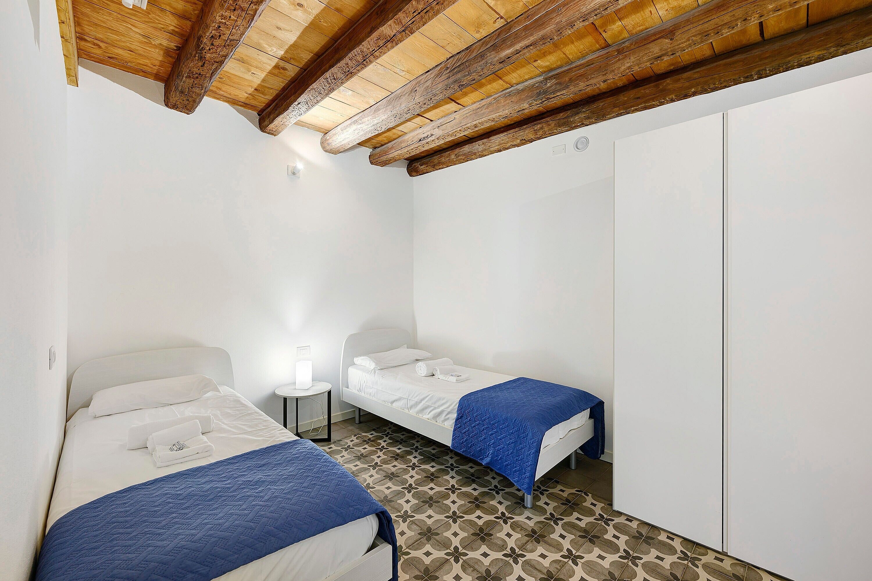 A room with two beds, a wardrobe, and a small table with a lamp.