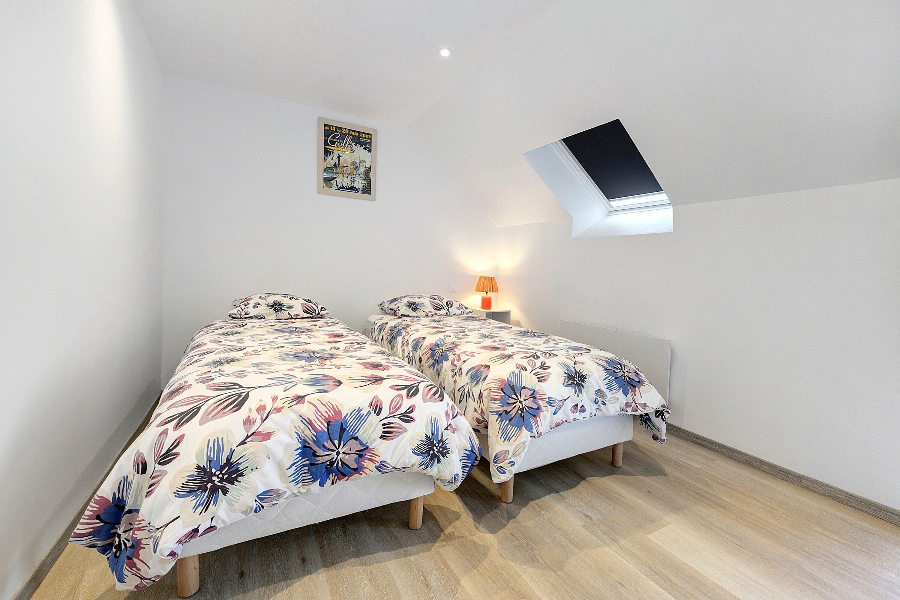 A bedroom with two beds, a skylight, a wall-mounted lamp, and a poster on the wall.