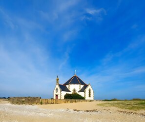 A white church with a black roof and a cross on top, surrounded by a sandy area with sparse vegetation.