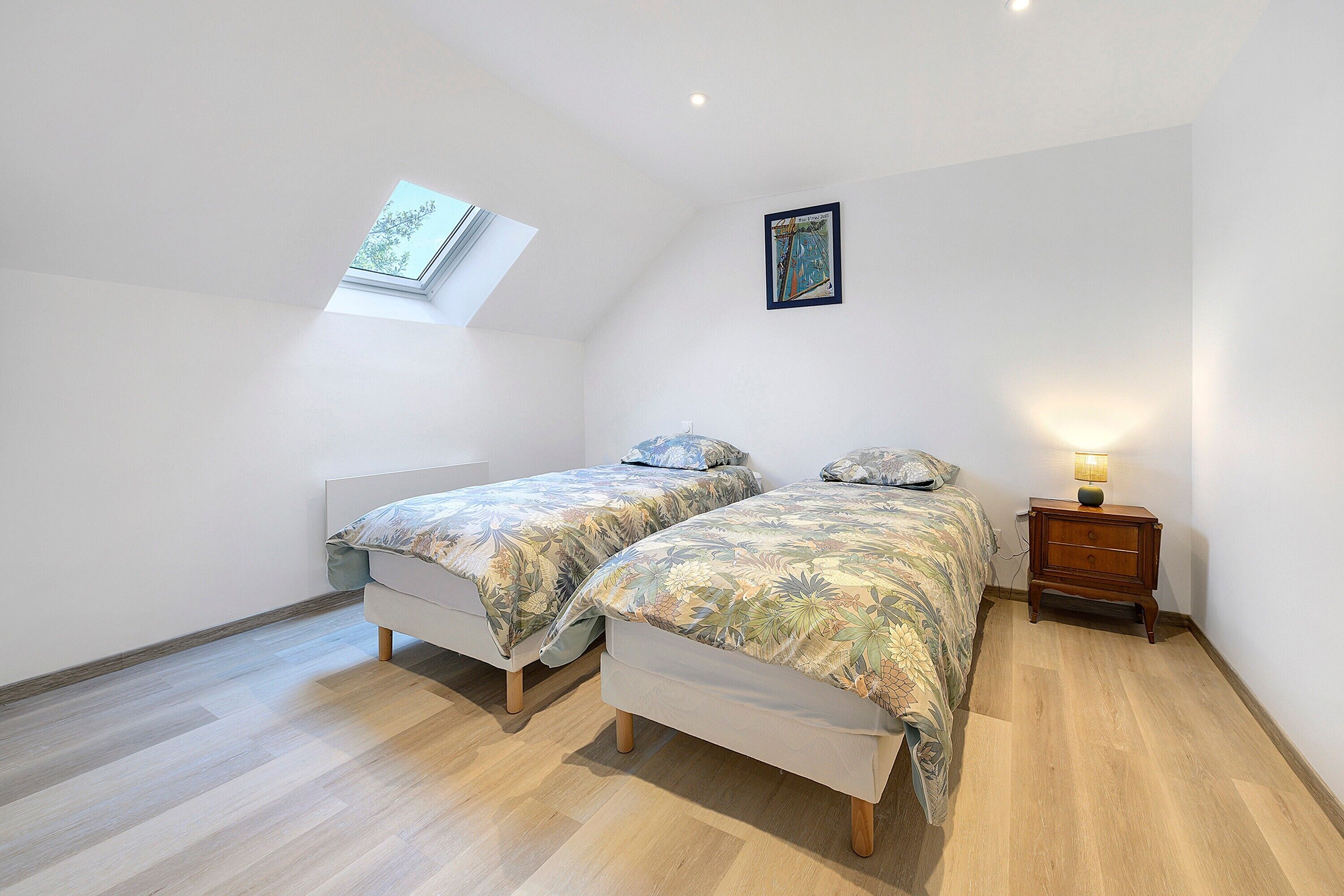 A bedroom with two beds, a skylight, a nightstand, and a framed picture on the wall.