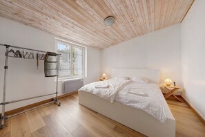 Ein Schlafzimmer mit Bett, Holzboden, einem Fenster, einem Nachttisch und einem Metallgestell mit Kleiderhängern.