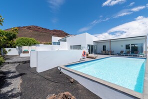 Un área de piscina moderna con una piscina azul cristalina, paredes blancas y una montaña al fondo.