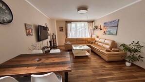 Ein Wohnzimmer mit einer Couch, einem Couchtisch, einem Esstisch mit Stühlen, einem an der Wand befestigten Fernseher und einer Pflanze.