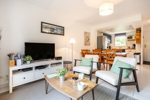 Ein modernes Wohnzimmer mit Flachbildfernseher, ein Essbereich mit Holzsesseln und eine durch eine offene Tür sichtbare Küche.