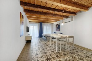 Ein Zimmer mit Holzbalkendecke, gemusterten Bodenfliesen, einem Esstisch mit Stühlen, einer Couch und einem Fernseher auf einem Schrank.