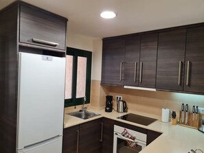 Une cuisine de taille réduite, équipée d’un réfrigérateur, d’un évier à deux bacs, d’une cuisinière et de meubles de rangement en bois foncé.
