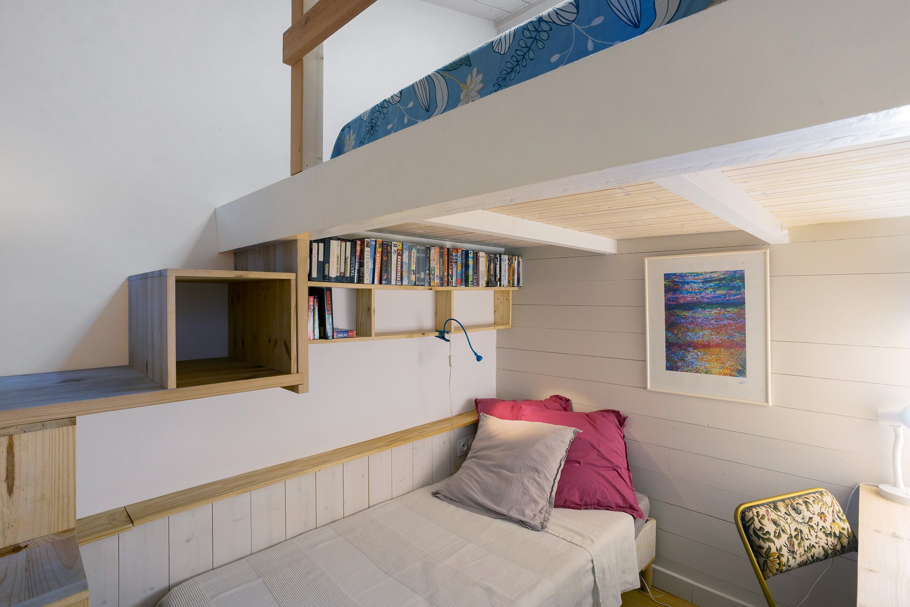 A bedroom with a bunk bed, a bookshelf, a bed with pillows, a chair, and a painting on the wall.