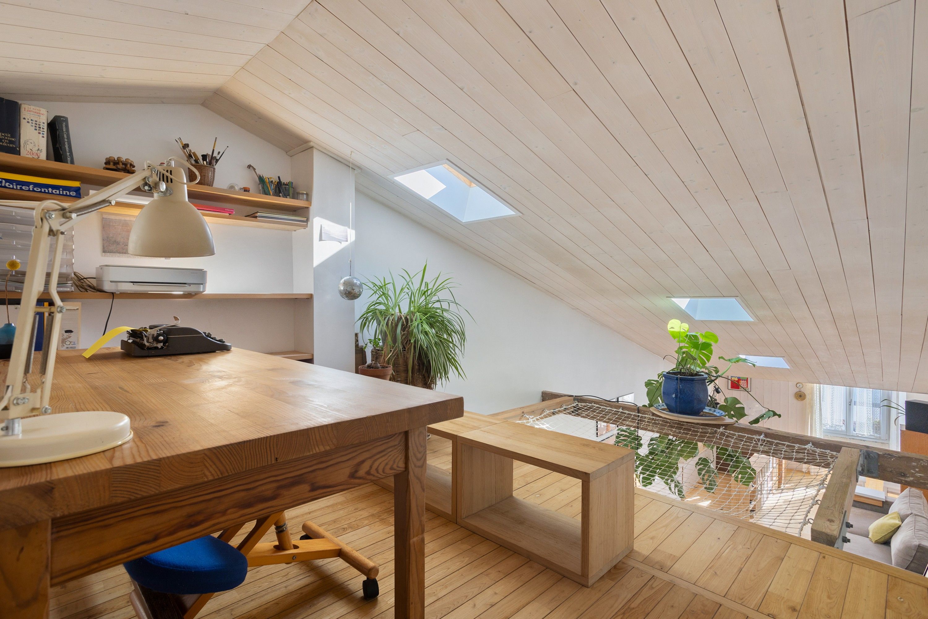 Habitación estilo loft con un escritorio de madera, una lámpara, una silla y un pequeño banco. Hay dos tragaluces y vistas al exterior a través de una ventana.