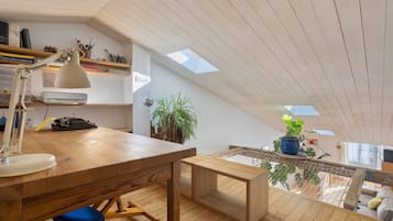 Habitación estilo loft con un escritorio de madera, una lámpara, una silla y un pequeño banco. Hay dos tragaluces y vistas al exterior a través de una ventana.