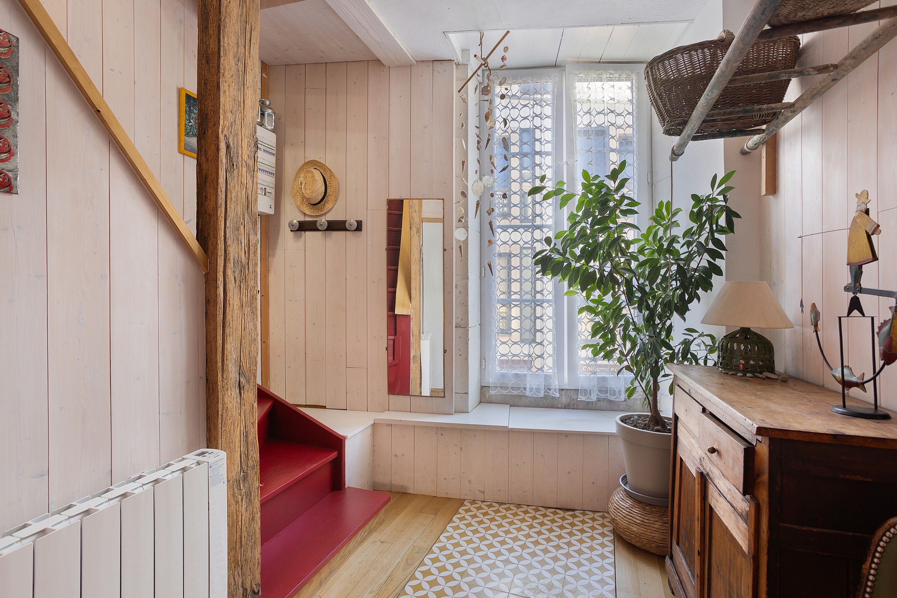 A cozy room with a wooden staircase, a red bench, a potted plant, and a window with a decorative screen.