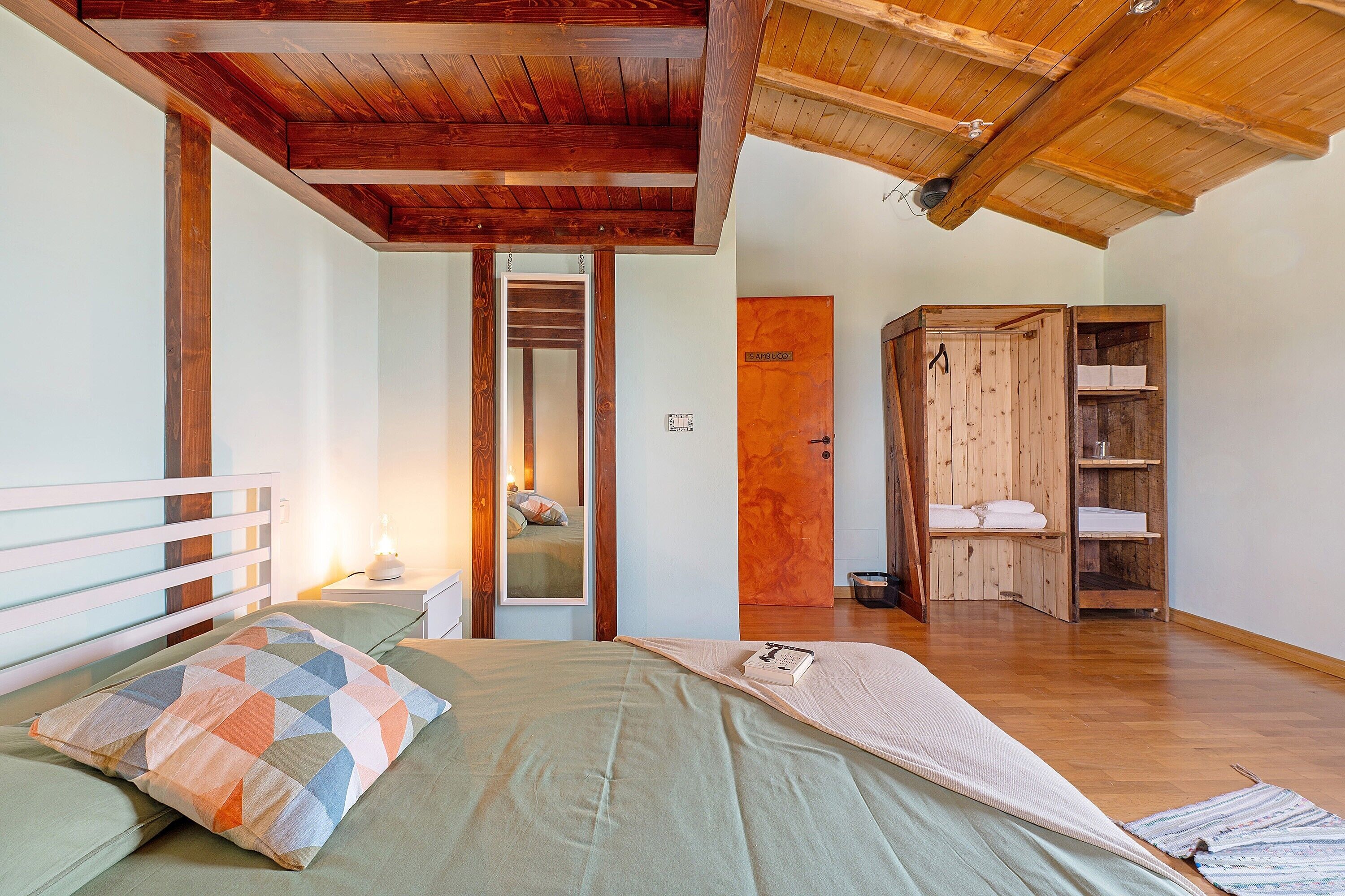 A bedroom with a wooden ceiling, a bed with a geometric-patterned pillow, a bedside table with a lamp, and a wooden wardrobe.