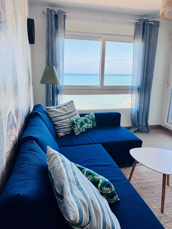 Quarto com sofá azul, mesa redonda pequena e vista para o mar através de grandes janelas.