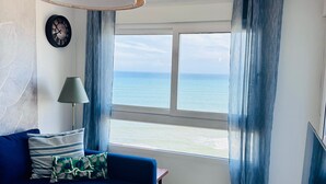 A room with a blue sofa, a small white table, a hanging lamp, a wooden side table, a clock, and a view of the sea through large windows.