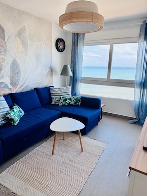 A living room with a blue sofa, a small white table, a wall clock, and a view of the sea through large windows.