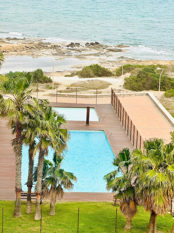 A pool with a wooden deck, surrounded by palm trees, overlooking a beach with rocks and a fence.