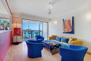 A modern living room with a yellow sofa, blue armchairs, a coffee table, and a large window offering a view of the ocean. - T3 Seafront Apartment with Sea View, Wi-Fi, and Air Conditioning (Cagnes-sur-Mer)