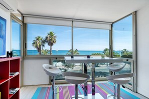 A modern room with a large window, a white table, two white bar stools, a red bookshelf, and a colorful rug. - T3 Seafront Apartment with Sea View, Wi-Fi, and Air Conditioning (Cagnes-sur-Mer)