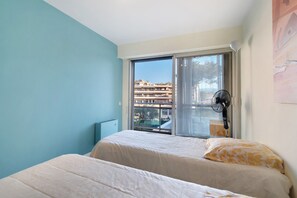 A bedroom with two beds, a window with a view of buildings, a fan, and a wall-mounted light. - T3 Seafront Apartment with Sea View, Wi-Fi, and Air Conditioning (Cagnes-sur-Mer)