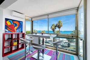 A modern room with a large window, a red bookshelf, a white table, and a colorful rug. - T3 Seafront Apartment with Sea View, Wi-Fi, and Air Conditioning (Cagnes-sur-Mer)