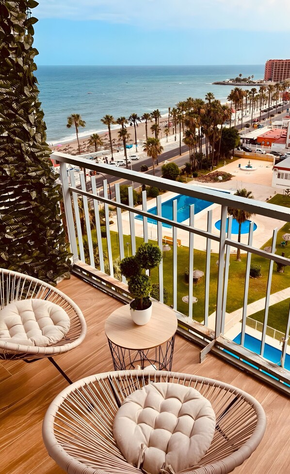 A balcony with wicker chairs, a small table with a plant, and a view of the ocean and beach.