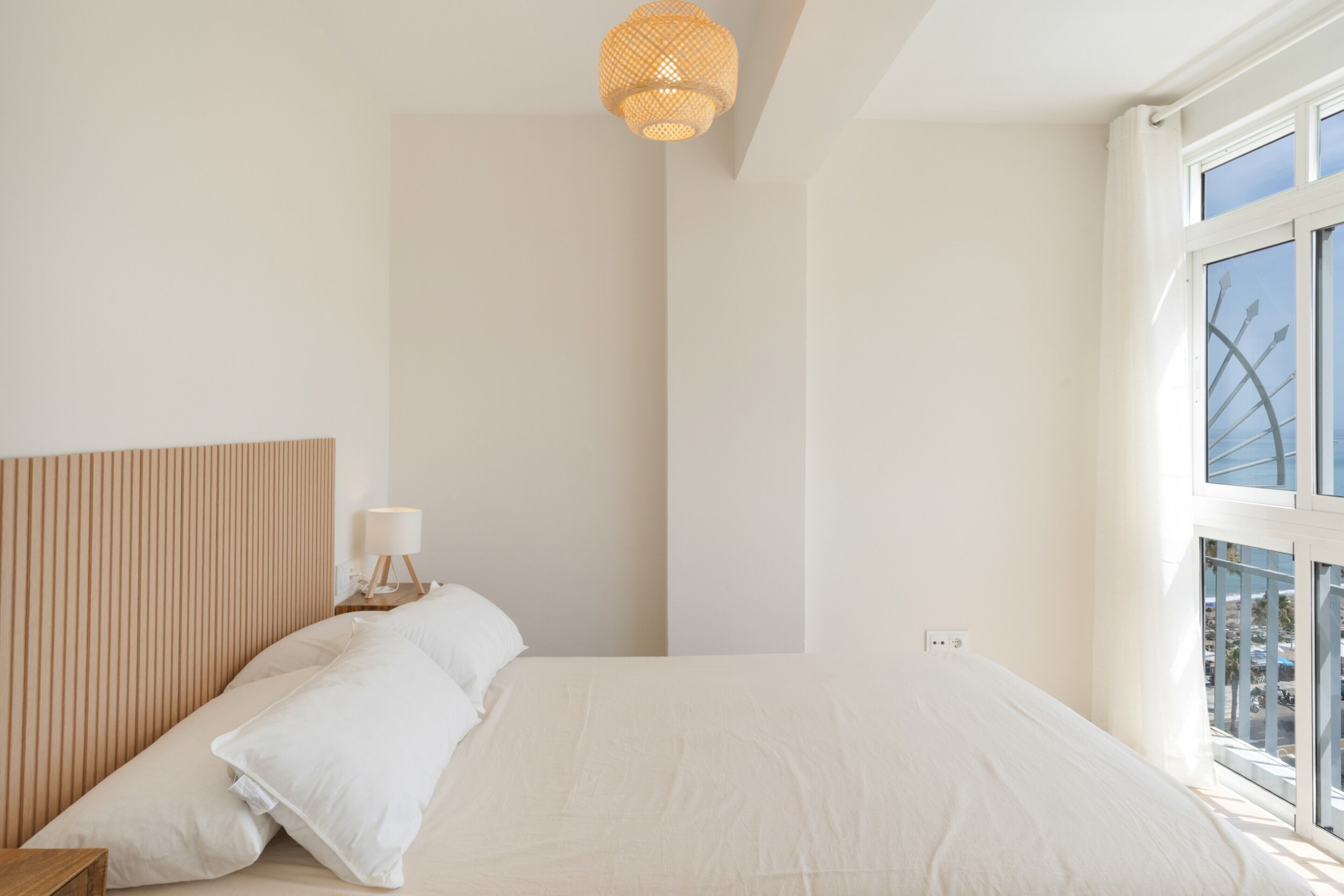 A minimalist bedroom with a large bed, a bedside table with a lamp, and a window with a view of buildings.