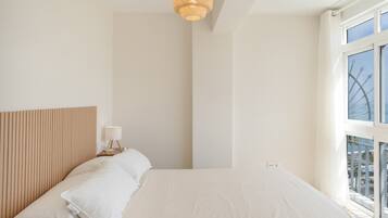 A minimalist bedroom with a large bed, a bedside table with a lamp, and a window with a view of buildings.
