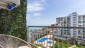 Ein Balkon mit Korbstuhl und kleinem Tisch, mit Blick auf ein Schwimmbecken und einen angelegten Garten.