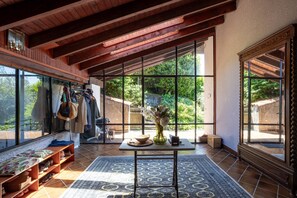 Habitación amplia con ventanas grandes, techo de madera, piso de baldosas y una mesa central con un florero y un cuenco.