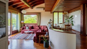 A spacious living area with a wooden ceiling, a red sofa, a dining table, and a kitchen area visible through an open door. - Groovy House: Vintage Charm, Lake Views, and Nature Retreat (Agra)
