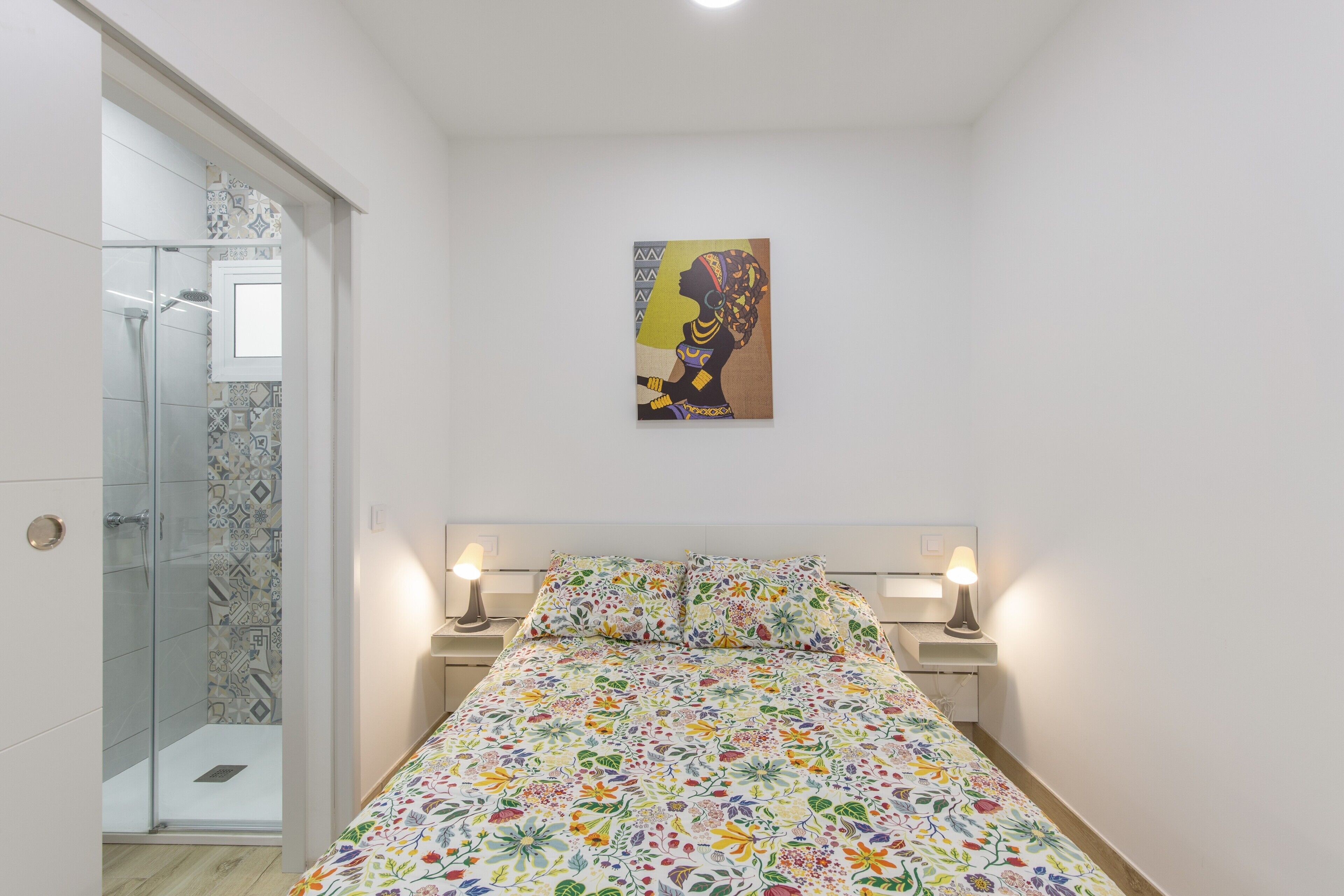 A bedroom with a bed, two bedside lamps, a glass shower enclosure, and a wall-mounted painting.