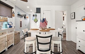 Ein Essbereich mit Tisch, Stühlen und amerikanischen Flaggen. Regale mit Küchenutensilien und ein Kühlschrank sind ebenfalls vorhanden.