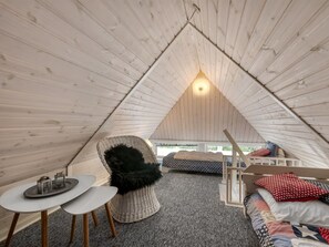 Ein gemütliches Schlafzimmer im Dachgeschoss mit schrägen Wänden, einem Korbstuhl, einem kleinen Tisch, einem Bett mit gemusterten Textilien und Blick ins Freie.