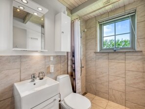 Ein Badezimmer mit weißem Waschbecken, Toilette und Dusche, einem Spiegel über dem Waschbecken und einem Fenster mit Blick auf Bäume.
